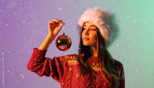Beautiful young woman wearing furry winter hat holding shiny golden christmas bauble indoors