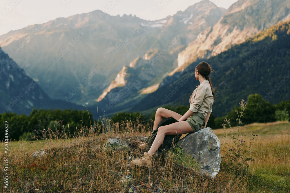 Obraz premium Woman sitting on a rock in a mountain meadow, enjoying outdoor scenery. Natural light highlights the valley and distant peaks, conveying serenity, exploration, and nature connection.