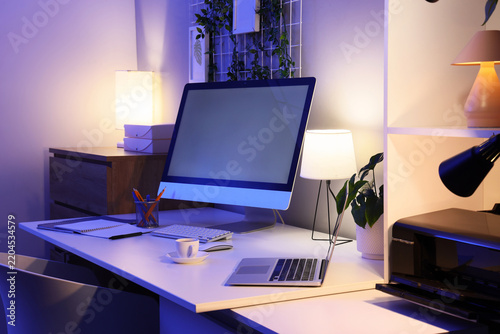 Workplace with blank computer monitor and glowing lamps in dark office