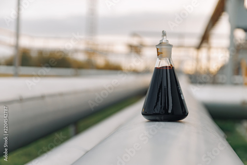 Dark petroleum sample in glass laboratory flask placed on greenhouse railing with blurred industrial structure and soft daylight in background, scientific oil research setting