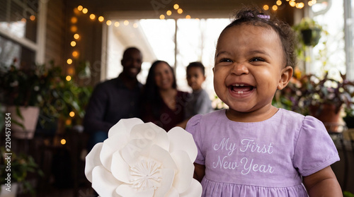 Delighted mixed-race baby girl in lavender My First New Year dress holding a paper flower laughs in a garden room, for springlike holiday greeting cards and posts.
