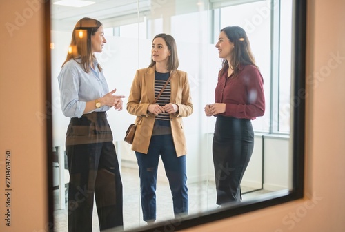 Three businesswomen in a professional setting discussing ideas. They appear to be in a modern office environment, engaged in a collegial conversation