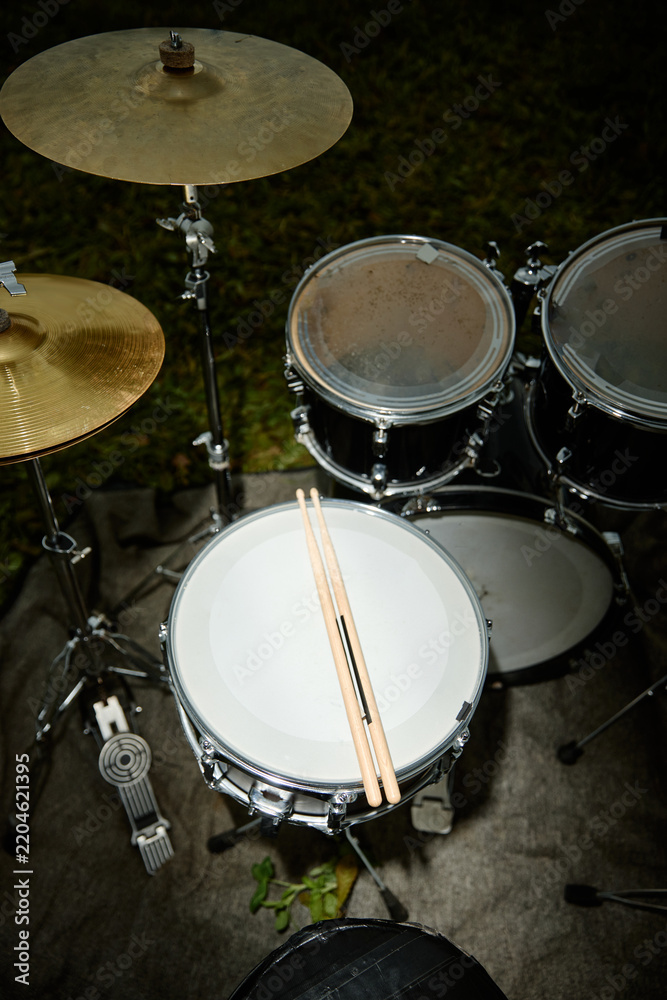 Obraz premium Vertical shot of drum kit with drumsticks resting on snare drum outdoors. Musical instrument set ready for live concert performance or indie music promotion