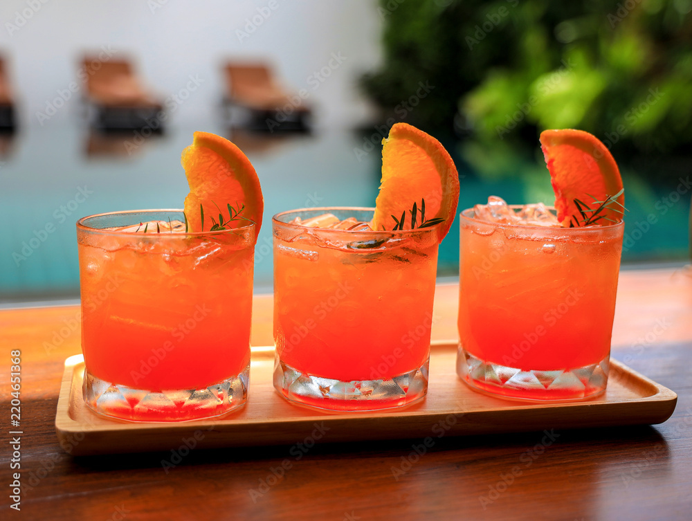 Obraz premium Three refreshing orange cocktails garnished with fresh orange slices and rosemary on a wooden tray by the poolside.