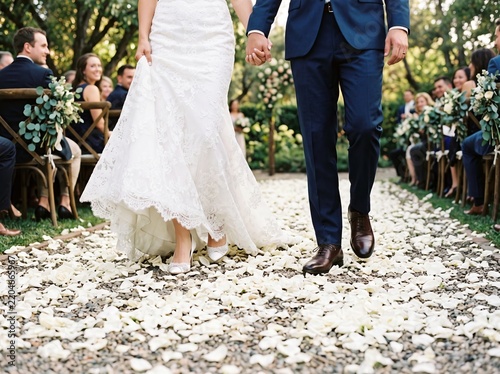 Young adult bride and groom holding hands while walking down a gravel wedding aisle covered in scattered white flower petals