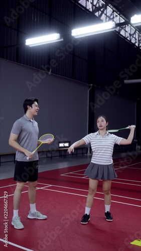 Asian male badminton coach guiding young woman through racket swing technique on indoor court