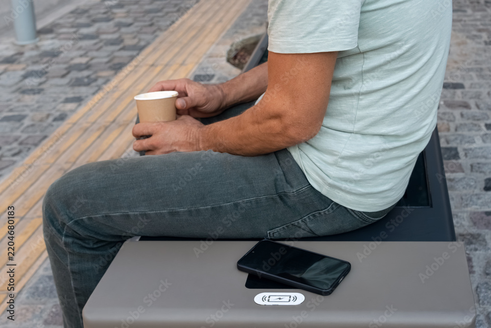 Fototapeta premium Man sitting on modern smart bench in city, holding coffee cup while his smartphone recharges on built-in wireless charging pad. Innovative urban infrastructure, street furniture, smart city lifestyle