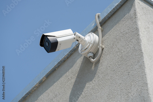 Outdoor bullet security CCTV camera mounted on a modern concrete building facade against a clear blue sky. Urban surveillance, private property protection, and building safety monitoring system.