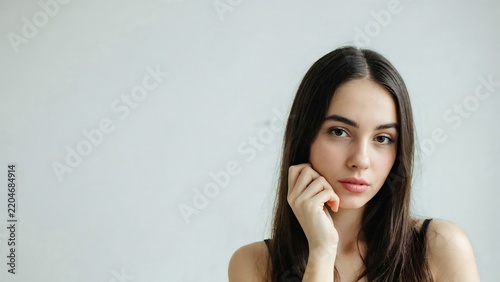 Portrait of a young woman with long dark hair resting her chin on her hand against a plain light gray wall