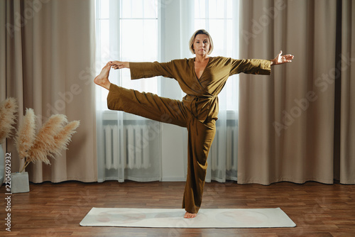 Middle-aged woman stands on one leg with her other leg bent and foot held in her hand. She spreads her arms wide as she focuses. Morning light streams through windows in calm indoor area.