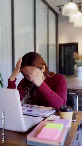 Stressed young Asian businesswoman holding head overwhelmed by paperwork at modern corporate office desk