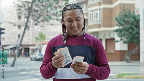 Fotografie Man smiling and counting pesos banknotes with hands on an urban street by apartment building and parked cars; satisfaction prosperity optimism
