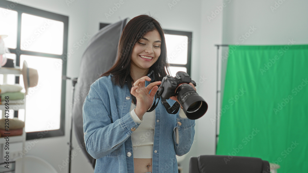 Obraz premium Young woman photographer adjusts a dslr camera and smiles while reviewing shots in a studio with green screen and softbox lighting; creative joy.