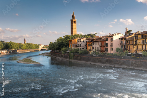 Cityscape of old city on river bank with sunlit roofs and bell tower on sunny day, Verona, Italy