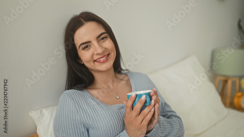Foto Woman smiling while holding a blue mug with both hands and bringing it to her lips on a bed with pillows and a wooden headboard in a bedroom; morning ritual serenity