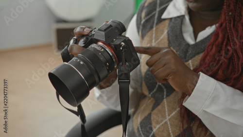 Woman points finger at camera while holding a mirrorless camera in a photo studio chair wearing argyle sweater vest and braided red hair; creative concentration.