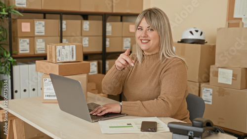 Woman points finger at camera while typing on laptop beside stacked parcels in a building; entrepreneurial confidence.
