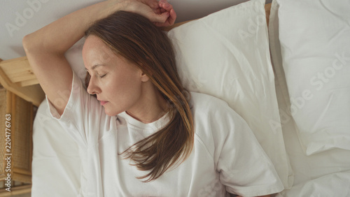 Woman lying in bed with arm above head and hand on stomach on white sheets beside a rattan nightstand in building; serenity rest.