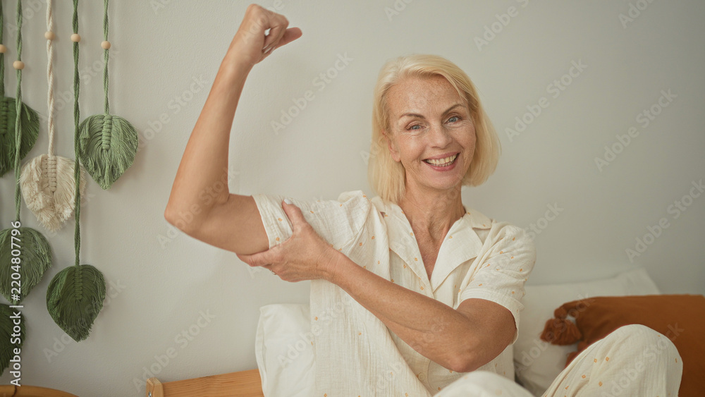 Obraz premium Woman flexes arm and holds bicep on a bed in a bright bedroom studio; confidence vitality wellness health.