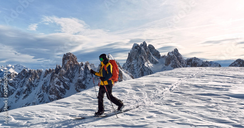Ski mountaineer crossing a snowy ridge in the Dolomites with dramatic alpine peaks and blue sky in the background, capturing winter adventure and breathtaking mountain landscape.