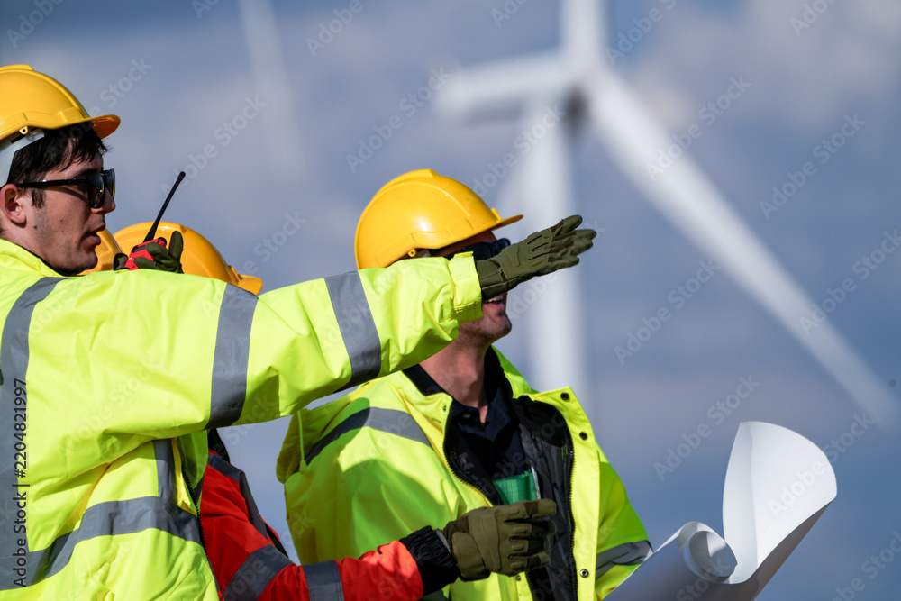 Fototapeta premium Two wind technicians with drawings shout observations over the rotor noise directly beneath a turning wind turbine in bright breezy daylight on a working wind farm in rural Thailand.