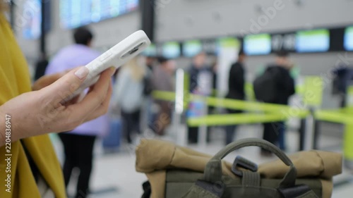 Woman holds phone, checks flight info at airport. Beside her, rolled backpack sits ready for travel. Background shows blurred travelers and check-in counters. Yellow tape separates queue areas