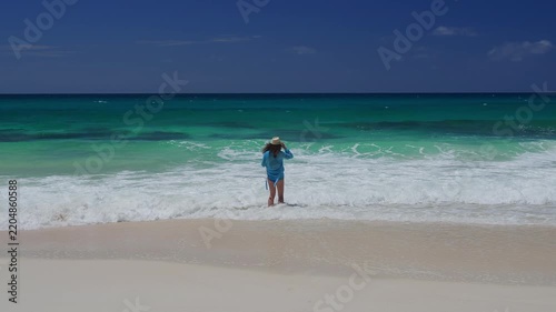 Person in blue rash guard enters turquoise surf beneath clear sky, static wide shot capturing coastal recreation moment with pristine sand, breaking waves, calm waters.