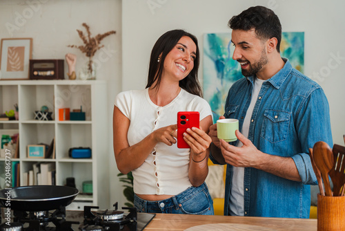 A couple engages in a light moment with a red smartphone while standing at a kitchen counter, sharing a quick glance and a cup of beverages. The scene conveys everyday connection and collaboration in