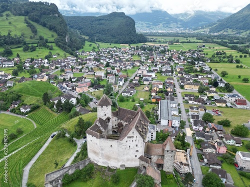 Aerial view of Gutenberg Castle surrounded by the village of Balzers, lush green hills, and mountains in Liechtenstein.