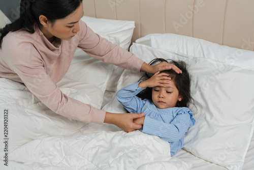 mother comforting sick daughter while touching her forehead with fever headache in bed