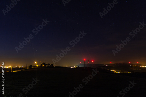 A horizontal photograph of a vast starry night sky over a dark rural landscape silhouette, featuring an illuminated windmill, distant city lights, and red wind turbine lights.