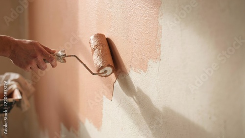 A hand painting a wall with a paint roller, applying a peach-colored coat to a textured surface