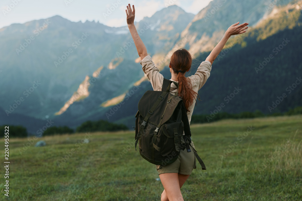 Obraz premium A woman with a backpack stands in a green meadow, arms raised toward distant mountains, capturing a moment of achievement, freedom, and outdoor adventure.
