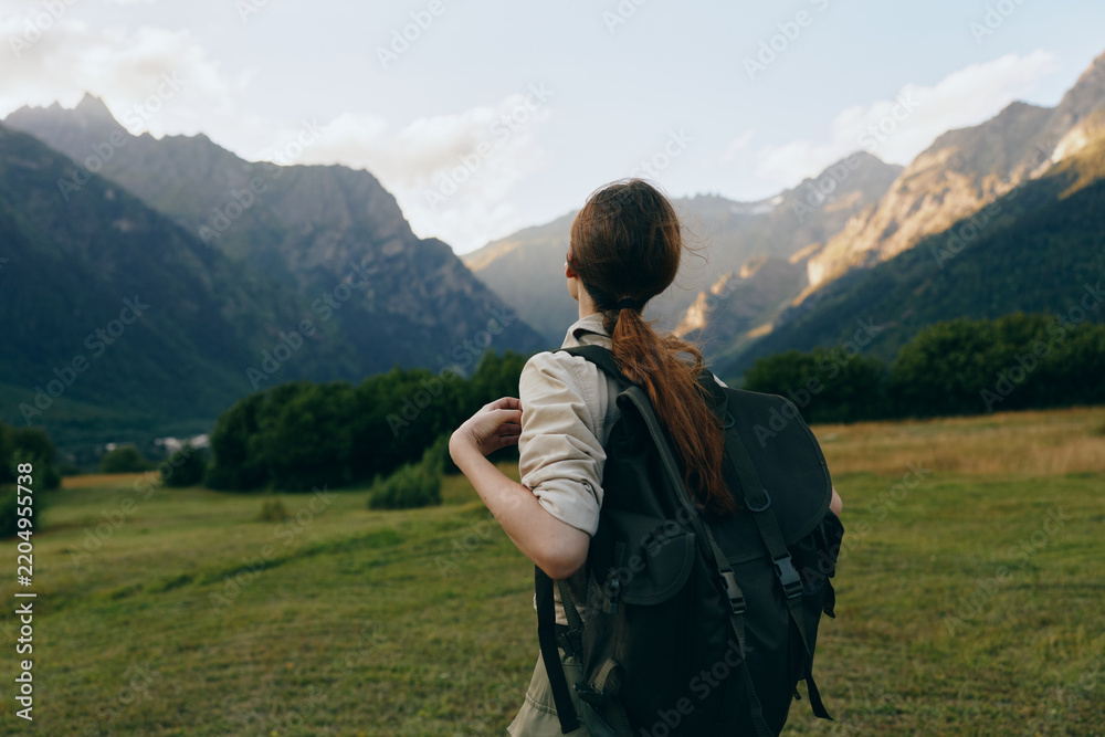 Obraz premium Woman with a backpack gazes over a mountain valley, exploring nature and outdoor life. Bright sky, green fields, distant peaks create a calm, adventurous mood for travel lifestyle.