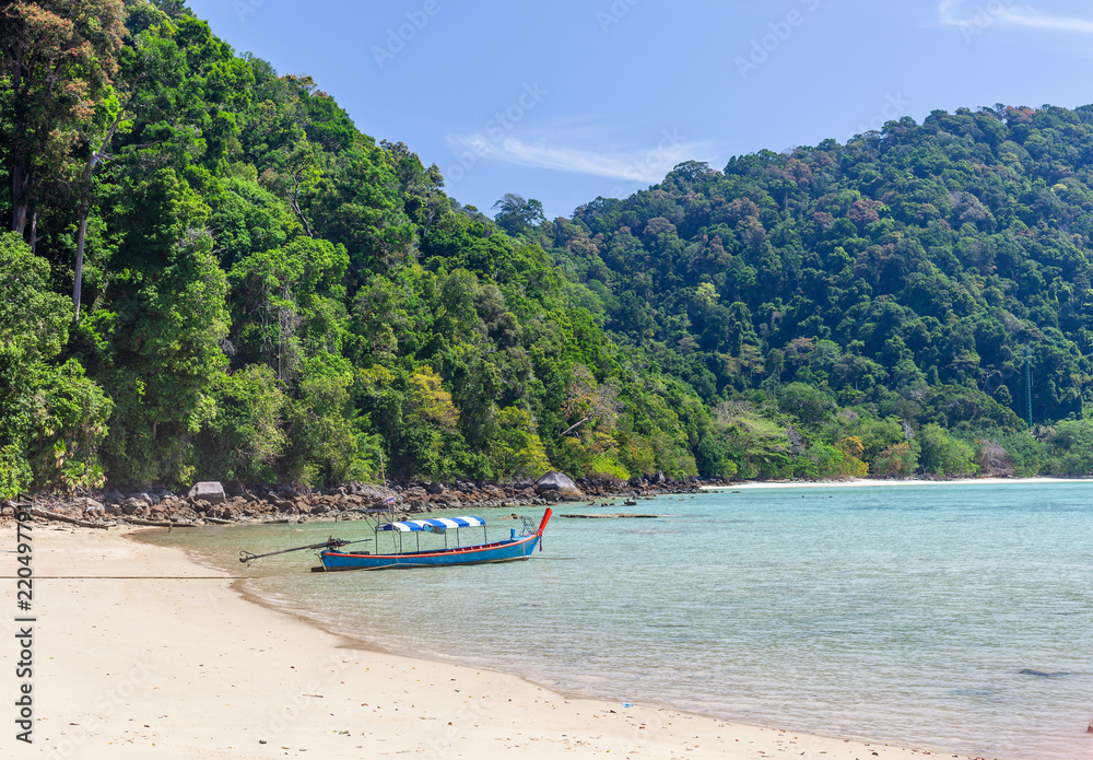 Fototapeta premium A longtail boat rests on a white sand beach beside clear turquoise water at Koh Surin, Thailand, with lush green hills under a blue sky.