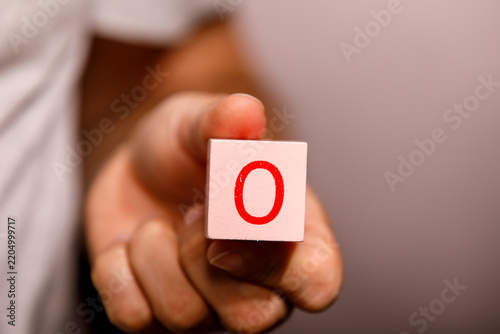 A hand extends forward, presenting a light pink wooden block with a red 'O' on it.