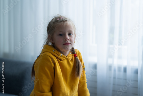 A girl sits on a sofa at home looking calmly into the camera, reflecting a mentally stable and secure state in a family setting. Child psychology articles, parenting resources, mental health