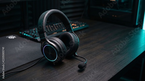Black gaming headset with microphone resting on a dark wooden desk in a dimly lit room with a keyboard in the background