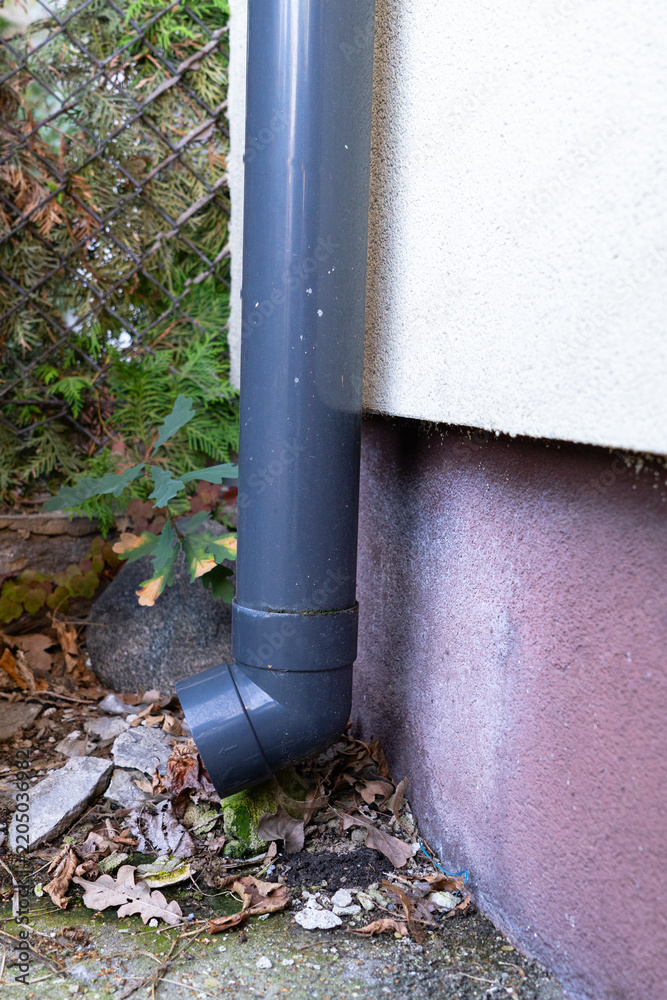Obraz premium Black rainwater downspout with elbow fitting beside a residential building, exterior drainage pipe connected to house gutter system