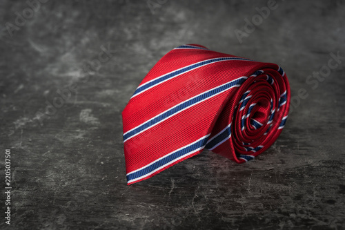 Elegant rolled red mens silk necktie with blue and white diagonal stripes on dark textured background for business fashion and executive dress code