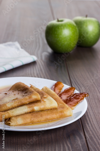 Crepes aka thin pancakes aka British pancakes with apple jam on the white plate on wooden table with green apples in background.