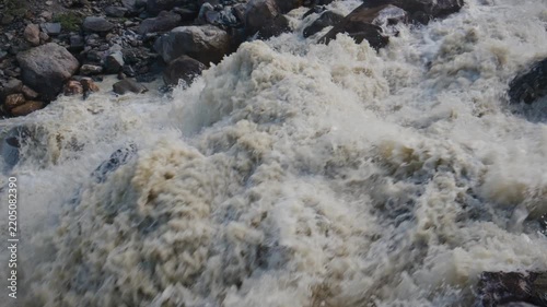 Raging river water crashes over dark rocks. Foam sprays upward in chaotic motion. Scene shows raw power of untamed nature. Ideal for adventure, environmental, or extreme weather themes