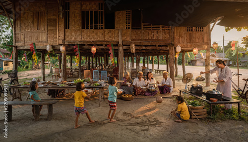 Children playing outside a traditional wooden stilt house with adults sitting nearby.