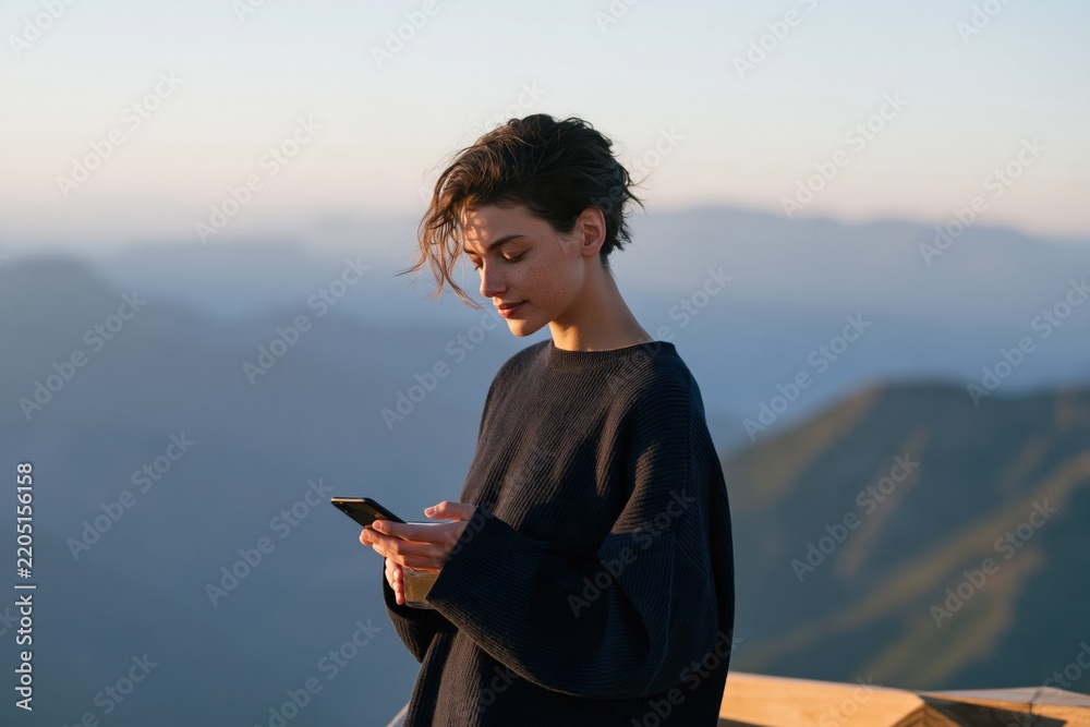 Naklejka premium Woman using smartphone on a mountain balcony at sunrise for travel lifestyle background and remote work communication