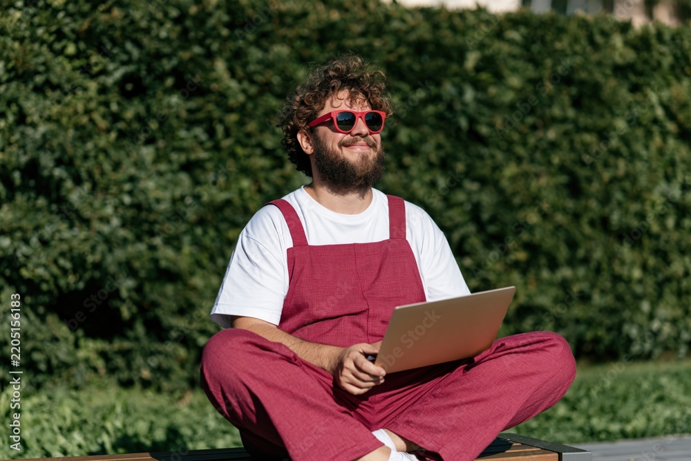 Fototapeta premium Curly haired man wearing red overalls using a laptop outdoors on a bench for remote work background and lifestyle banner design