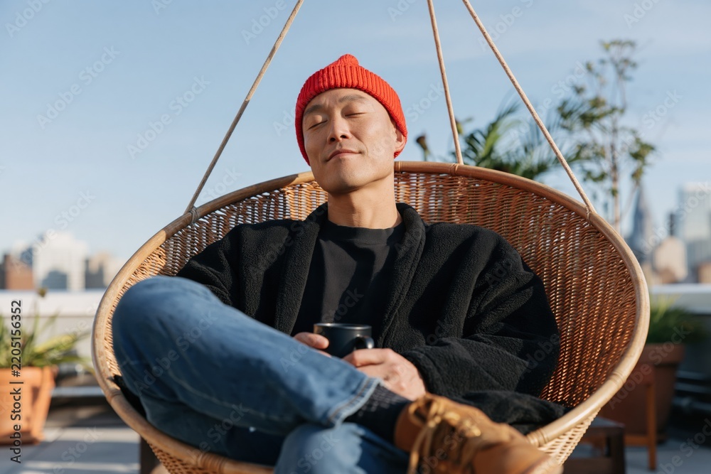 Fototapeta premium Rooftop relaxing man in red beanie sitting in a hanging wicker chair with coffee mug for lifestyle background and urban leisure design