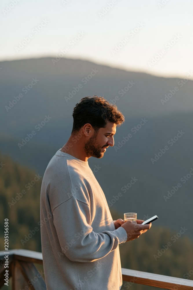 Fototapeta premium Man on mountain balcony checking a smartphone while holding a glass of drink at sunset for travel lifestyle background and evening relaxation