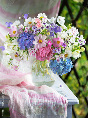Beautiful bouquet of flowers in a vase on a table—phlox, snapdragons, hydrangeas, and bellflowers—on a terrace against the backdrop of a pergola and garden, blur, selective focus.