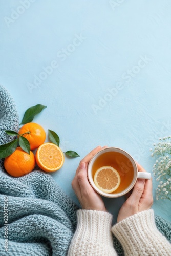 Hands holding cup of lemon tea with fresh oranges on knitted blanket on light blue background for winter immunity