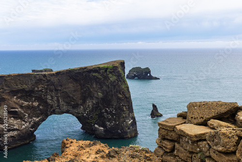 Dyrholaey Arch rises above the Atlantic Ocean in South Iceland, with rugged volcanic sea cliffs, offshore rock formations and an expansive cloudy coastal horizon.